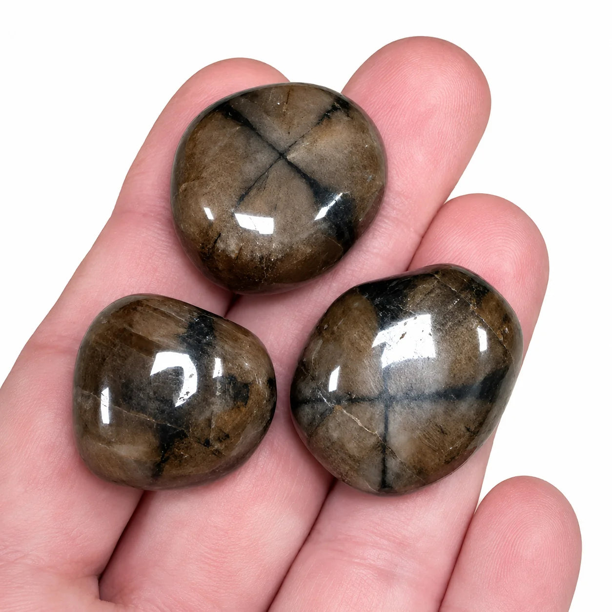 Trois pierres naturelles andalousite variété chiastolite roulées de Chine présentées dans une main pour illustrer leur taille réelle d'environ 2 à 3 cm, pierres polies aux croix naturelles bien marquées.